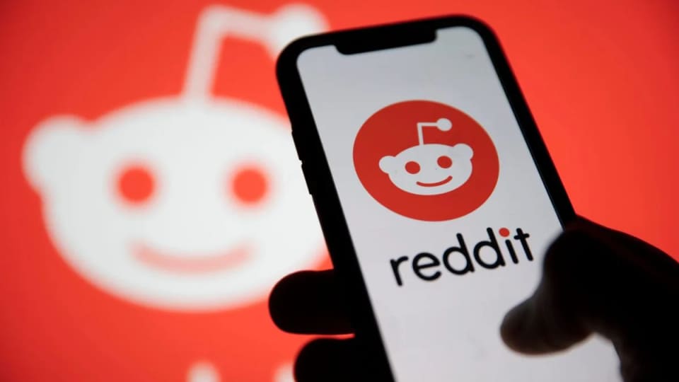 A hand holding a smartphone displaying the Reddit app logo against a red Reddit background.