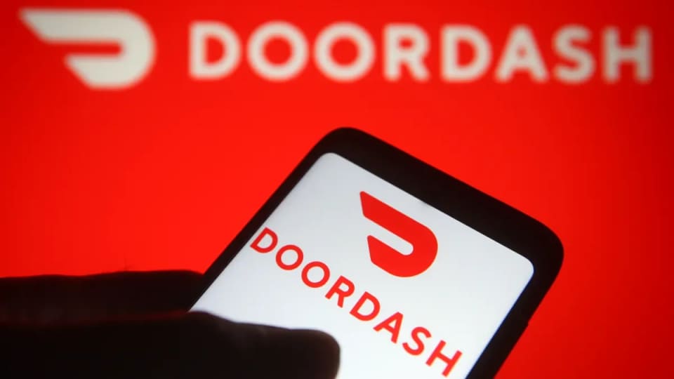 A hand holding a phone displaying the DoorDash app logo against a red DoorDash sign.